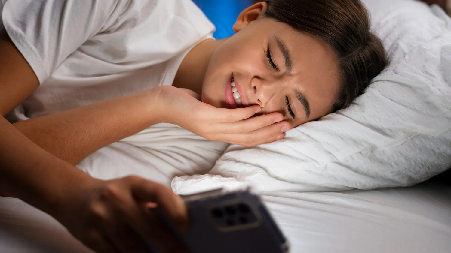 A tired woman in bed checks her phone, getting ready for interview calls while still feeling sleepy and cozy.